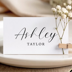 Könnte beinhalten: Eine rechteckige weiße Platzkarte mit dem Namen Ashley in schwarzer Schreibschrift und dem Namen TAYLOR in einer serifenlosen schwarzen Schrift. Eine kleine Holzklammer befestigt einen Zweig weißer Schleierkrautblüten an der rechten Seite.