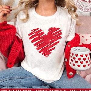 May include: A white t-shirt with a red, hand-drawn heart design. The shirt is being worn by a person with a red cardigan and blue jeans. The text "Love" is on a white mug with a red heart pattern.