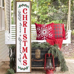 Può includere: Un'insegna in legno a tema natalizio con la parola "CHRISTMAS" in lettere maiuscole rosse e "have yourself a merry little" in corsivo. L'insegna è decorata con agrifoglio. Una lanterna rossa e cuscini sono sullo sfondo.