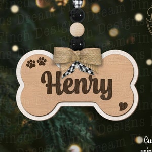May include: A wooden dog bone ornament with the name "Henry" engraved in a dark brown font. The ornament features paw prints, a heart, and a burlap bow with a black and white checkered accent. It hangs from a string of black and wooden beads.