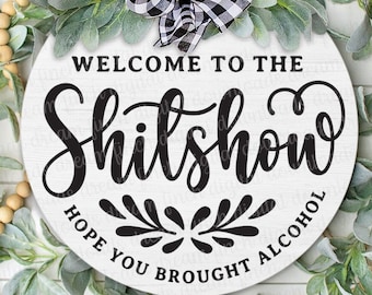 May include: A white round wooden sign with the text "Welcome to the Shitshow Hope you brought alcohol" in black cursive lettering. The sign is decorated with a black and white checkered bow and a garland of green leaves and wooden beads.
