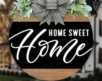Könnte beinhalten: Ein rundes Holzschild mit schwarzem Hintergrund und weißem Text, der "Home Sweet Home" lautet. Das Schild ist mit einer schwarz-weißen Schleife und Grünpflanzen verziert.