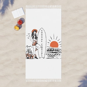 Puede incluir: Toalla de playa blanca con una ilustración en negro y naranja de una mujer con una tabla de surf, el texto "COASTAL COWGIRL", una puesta de sol y olas del mar. La toalla tiene un borde con flecos.