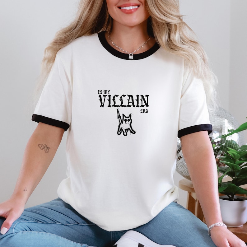 Pu&ograve; includere: T-shirt color crema con bordi neri sul colletto e sulle maniche. La maglietta presenta la scritta "IN MY VILLAIN ERA" con un'illustrazione di un gatto in nero. La maglietta &egrave; realizzata in un materiale morbido.