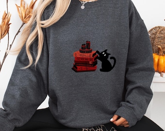 Op de afbeelding: Donkergrijze sweatshirt met een zwarte kat die naar een stapel rode boeken met kaarsen reikt. Het ontwerp heeft een Halloween- of heksenthema, perfect voor de herfst.
