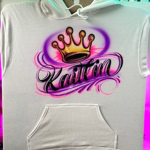 May include: White hoodie with a purple and pink airbrushed design featuring a gold crown and the name "Kaitlin" in cursive.