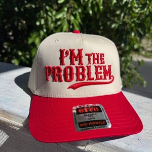 Op de afbeelding: Een beige en rode baseballcap met de geborduurde tekst "I'M THE PROBLEM". De cap heeft een rode klep en een rode knop bovenop. De cap is een klassieke pasvorm, mid profile en OSFM.