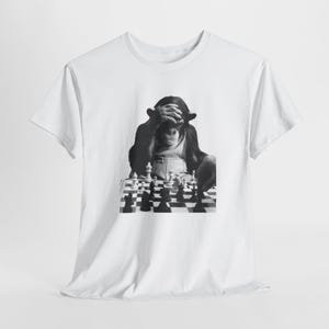Può includere: T-shirt bianco con un'immagine in bianco e nero di uno scimpanzé che gioca a scacchi, con le mani sulla testa. La scacchiera è a fuoco, con i pezzi disposti sulla scacchiera a scacchi. La maglietta è realizzata in un materiale morbido.