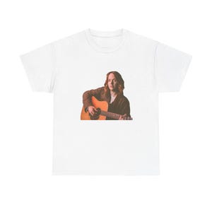 May include: White t-shirt featuring a person playing an acoustic guitar. The person is wearing a brown jacket and is positioned on the right side of the shirt. The guitar is a light brown color.