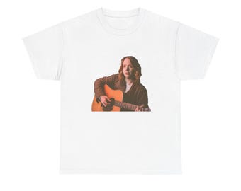 May include: White t-shirt featuring a person playing an acoustic guitar. The person is wearing a brown jacket and is positioned on the right side of the shirt. The guitar is a light brown color.