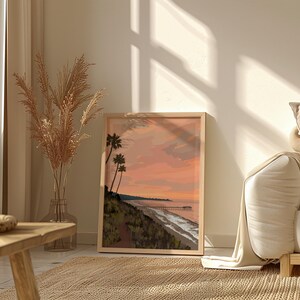 May include: A framed print of a sunset over a beach with palm trees in the foreground. The print is in a light wood frame and is hanging on a white wall.