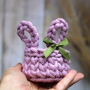 Könnte beinhalten: Ein kleiner, rosa gehäkelter Korb mit Hasenohren und einer grünen Schleife.