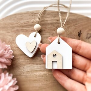 Peut inclure: Deux ornements en bois blancs, l'un en forme de c&oelig;ur et l'autre en forme de maison, chacun avec un petit accent en bois. Les deux sont suspendus par de la ficelle et des perles en bois. Le c&oelig;ur a un petit c&oelig;ur, et la maison a une d&eacute;coupe en forme de maison.