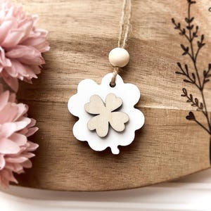 Peut inclure: Un porte-bonheur en forme de tr&egrave;fle en bois blanc avec un tr&egrave;fle en bois naturel au centre, suspendu &agrave; une ficelle. Le charme est pos&eacute; sur un fond en bois avec des fleurs roses &agrave; gauche.