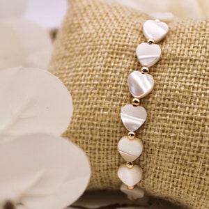 Könnte beinhalten: Ein weißes Armband mit herzförmigen Perlen und goldenen Akzenten. Das Armband wird auf einer braunen Jutestoff-Oberfläche präsentiert.