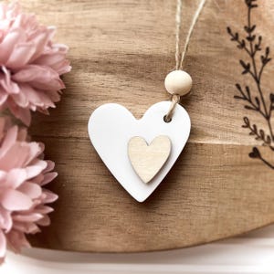 Peut inclure: Ornement en bois en forme de c&oelig;ur blanc avec un petit c&oelig;ur au centre, suspendu par de la ficelle et une perle en bois. L'ornement est pos&eacute; sur un fond en bois avec des fleurs roses &agrave; gauche.