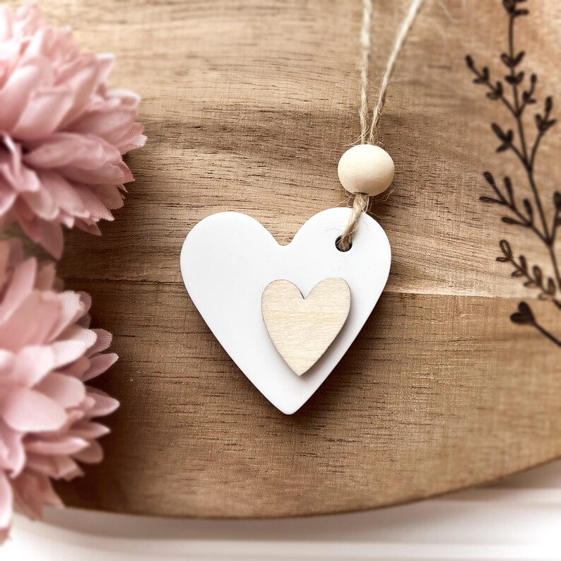 Peut inclure: Ornement en bois en forme de c&oelig;ur blanc avec un petit c&oelig;ur au centre, suspendu par de la ficelle et une perle en bois. L'ornement est pos&eacute; sur un fond en bois avec des fleurs roses &agrave; gauche.