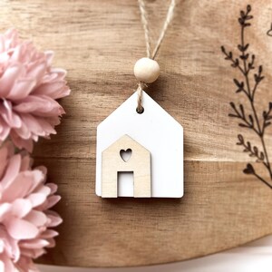 Peut inclure: Ornement en forme de maison en bois blanc avec une petite maison d&eacute;coup&eacute;e en forme de c&oelig;ur. L'ornement est suspendu &agrave; une ficelle en ficelle naturelle avec une perle en bois. Le fond est une surface en bois avec des fleurs roses.
