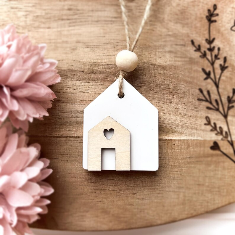 Peut inclure: Ornement en forme de maison en bois blanc avec une petite maison d&eacute;coup&eacute;e en forme de c&oelig;ur. L'ornement est suspendu &agrave; une ficelle en ficelle naturelle avec une perle en bois. Le fond est une surface en bois avec des fleurs roses.