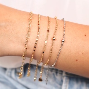 Könnte beinhalten: Ein Set aus fünf goldenen Kettenarmbändern in verschiedenen Stilen, darunter eine herzförmige Kette, eine zarte Kette und eine Kette mit kleinen Perlen. Die Armbänder sind auf dem Handgelenk einer Person übereinander gelegt.