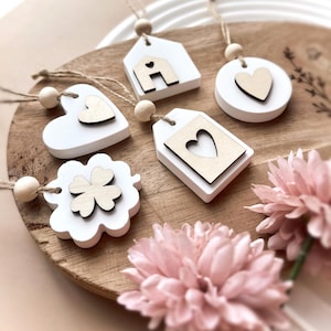 Può includere: Una collezione di ornamenti in legno bianco con dettagli in legno naturale, tra cui una casa, un cuore, un'etichetta, un cerchio e un quadrifoglio. Ogni ornamento ha un cordino per appenderlo e una perlina di legno. Gli ornamenti sono esposti su una superficie di legno con fiori rosa.