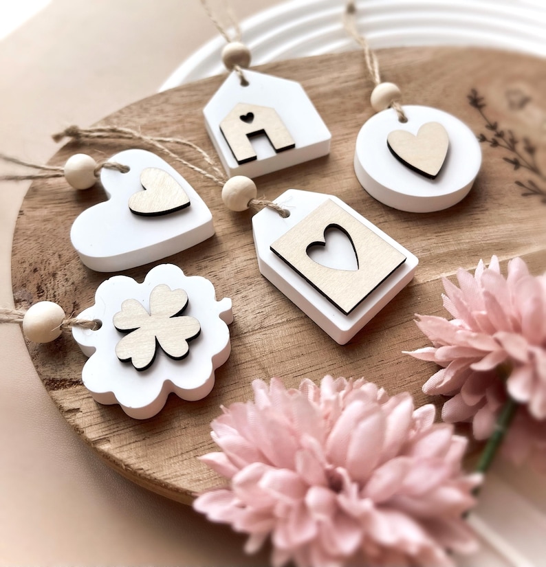 Peut inclure: Une collection d'ornements en bois blanc avec des d&eacute;tails en bois naturel, dont une maison, un c&oelig;ur, une &eacute;tiquette, un cercle et un tr&egrave;fle &agrave; quatre feuilles. Chaque ornement a une ficelle pour suspendre et une perle en bois. Les ornements sont pr&eacute;sent&eacute;s sur une surface en bois avec des fleurs roses.