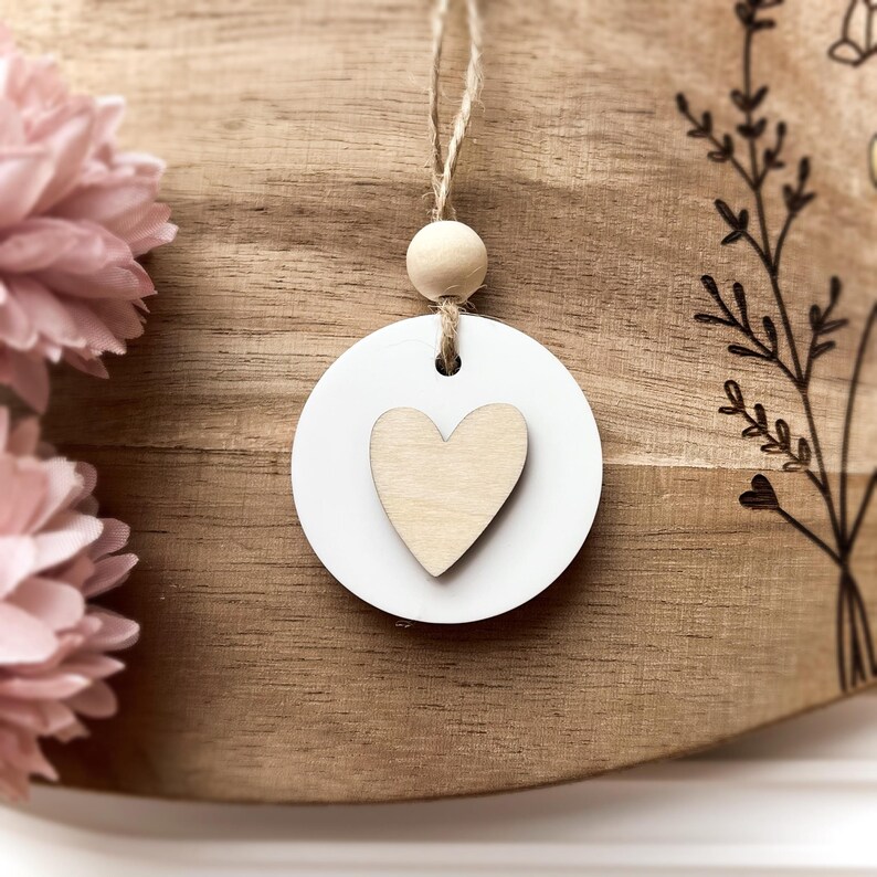 Peut inclure: Un ornement circulaire blanc avec un c&oelig;ur en bois, suspendu par de la ficelle et une perle en bois. L'ornement est pos&eacute; sur un fond en bois avec des motifs floraux et des fleurs en tissu rose.