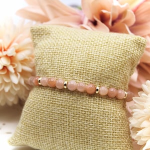 Op de afbeelding: Een delicate armband gemaakt met kleine, lichtroze en perzikkleurige kralen gescheiden door gouden spacers. De armband wordt getoond op een lichtbruin stofoppervlak.