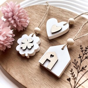 Peut inclure: Trois ornements en bois blancs avec des accents en bois naturel et des cintres en ficelle. L'un est un tr&egrave;fle &agrave; quatre feuilles, un autre un c&oelig;ur et le troisi&egrave;me une forme de maison. Une perle en bois est au sommet de chaque cintre. Des fleurs roses sont en arri&egrave;re-plan.