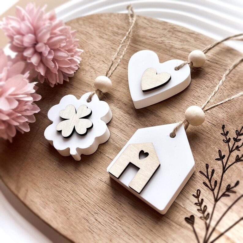 Peut inclure: Trois ornements en bois blancs avec des accents en bois naturel et des cintres en ficelle. L'un est un tr&egrave;fle &agrave; quatre feuilles, un autre un c&oelig;ur et le troisi&egrave;me une forme de maison. Une perle en bois est au sommet de chaque cintre. Des fleurs roses sont en arri&egrave;re-plan.