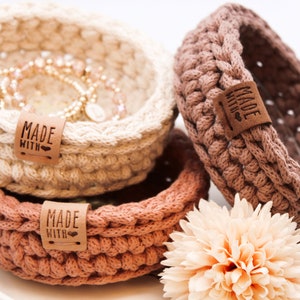 Könnte beinhalten: Drei gehäkelte Körbe in Beige, Hellbraun und Dunkelbraun mit einem Lederanhänger, auf dem "Made with Love" steht. Die Körbe sind übereinander gestapelt und eine hellpfirsichfarbene Blume befindet sich im Vordergrund.