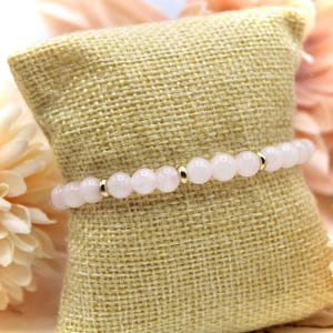 Könnte beinhalten: Ein Armband aus rosa Rosenquarz mit goldenen Perlen. Das Armband liegt auf einer beigefarbenen Stoffoberfläche.