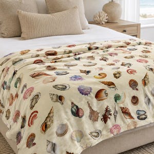 Puede incluir: Una manta de color crema con un patrón repetitivo de varias conchas marinas en tonos marrón, bronceado, rosa, morado y azul. La manta está colocada sobre una cama, mostrando una colección diversa de caracolas, vieiras y almejas.