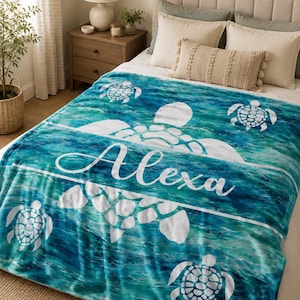 May include: A soft throw blanket featuring a vibrant blue and teal ocean water pattern with white sea turtle silhouettes and the name Alexa printed in a white script font across the centre of the blanket, covering the bed surface.