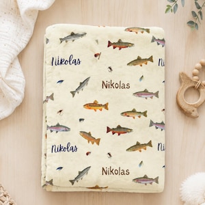 May include: A cream-colored fleece blanket featuring a repeating pattern of various colorful freshwater fish and fishing lures, interspersed with the name Nikolas printed in dark blue and brown text throughout the fabric surface.