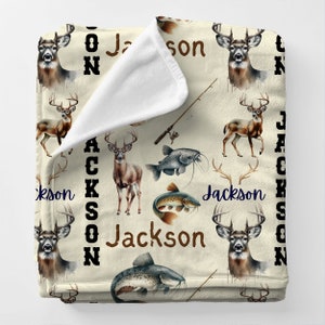 Puede incluir: Una manta de forro polar de color crema con un patrón repetido de ciervos marrones, cañas de pescar y bagres. El nombre "Jackson" se repite en todo el patrón.