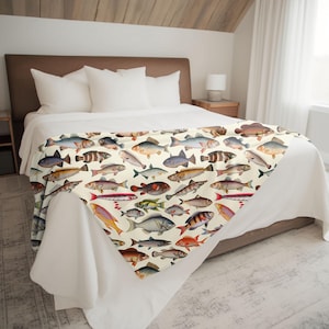 May include: A white throw blanket with a colorful pattern of various fish illustrations. The blanket is folded and displayed on a bed with white bedding.