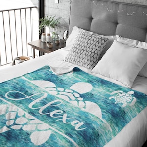 May include: A teal and blue fleece blanket with a white sea turtle design and the name "Alexa" printed on it. The blanket is on a bed with white sheets and gray pillows.
