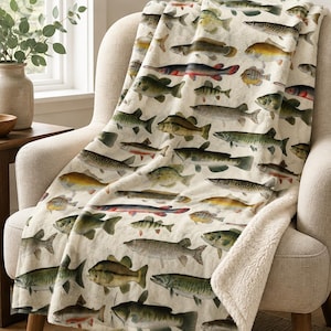 May include: A plush throw blanket featuring a repeating pattern of various freshwater fish species in shades of green, brown, and silver on a cream background. The blanket has a soft, textured white sherpa fleece lining visible along the folded edge.