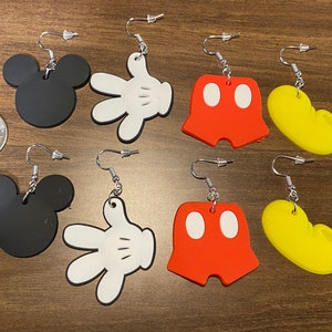 Peut inclure: Six paires de boucles d'oreilles pendantes repr&eacute;sentant diff&eacute;rents personnages embl&eacute;matiques de Mickey Mouse. Les boucles d'oreilles sont en plastique noir, blanc, rouge et jaune. Chaque paire repr&eacute;sente un personnage diff&eacute;rent&nbsp;: la t&ecirc;te de Mickey Mouse, le gant de Mickey Mouse, le short de Mickey Mouse et les chaussures de Mickey Mouse.