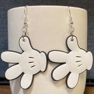 Peut inclure: Une paire de boucles d'oreilles en forme de gant de Mickey Mouse blanc avec des contours noirs et des crochets argent&eacute;s.