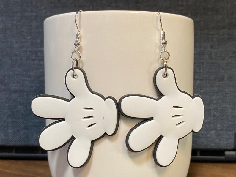 Peut inclure: Une paire de boucles d'oreilles en forme de gant de Mickey Mouse blanc avec des contours noirs et des crochets argent&eacute;s.