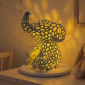 May include: A decorative elephant-shaped lamp with a textured, patterned design. The lamp emits a warm, yellow glow, casting light through its openings. It sits on a white surface, next to a stuffed bunny and books, in a nursery setting.