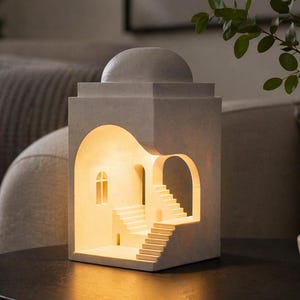 Könnte beinhalten: Eine graue Lampe im architektonischen Stil mit einem gewölbten Dach und ausgeschnittenen Bögen und Treppen. Die Lampe strahlt ein warmes Licht aus und schafft eine gemütliche Atmosphäre. Die Lampe steht auf einem dunklen Tisch, mit einer dekorativen Schale und einer Pflanze im Hintergrund.