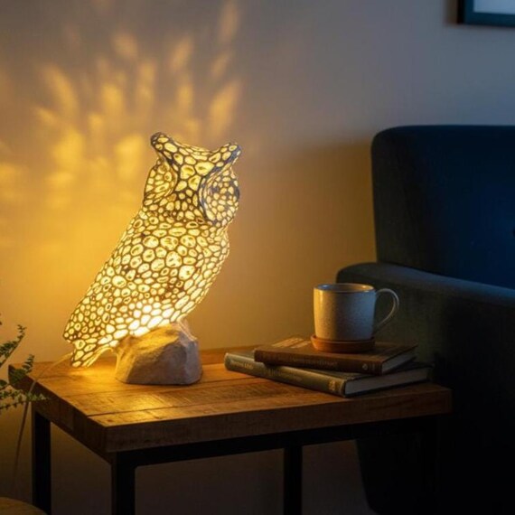 May include: A decorative owl-shaped table lamp. The lamp is white with a textured pattern and sits on a stone base. The lamp is on a wooden table with books and a mug. The lamp emits a warm, inviting glow.