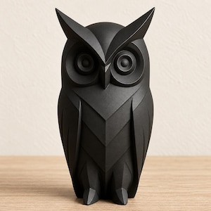 May include: A matte black owl figurine featuring a geometric, faceted design. The owl has pointed ear tufts, large circular eyes, and a sharp beak, with angular wing and chest details that create a structured, layered appearance throughout the body.