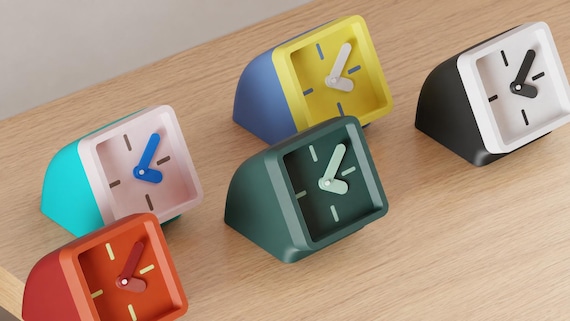 Angled close-up of Framelean Desk Clock examples in contrasting colors on light oak against a gray wall.