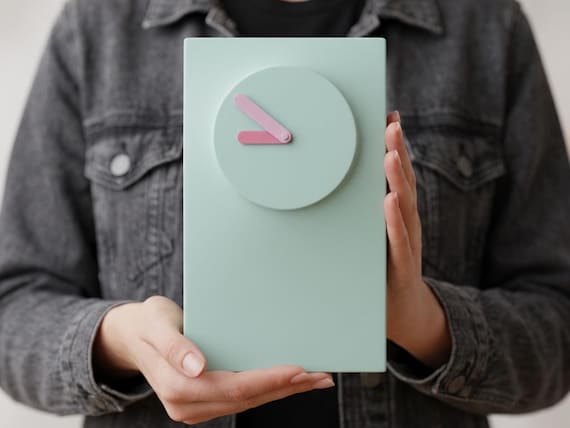 Softline Wall and Desk Clock held upright by a person, showing its pale blue rectangular body, raised round dial and pink hands.