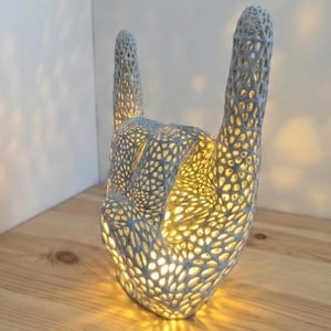 May include: A light blue, openwork hand sculpture in the 'rock on' gesture, illuminated from within. The intricate design features a pattern of small, irregular holes, creating a warm glow. The lamp sits on a wooden surface.