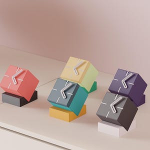 May include: A collection of cube-shaped clocks in various colors including red, yellow, teal, purple, and dark gray. Each clock features white hands and simple white markings on the face, resting on small rectangular bases of contrasting colors.